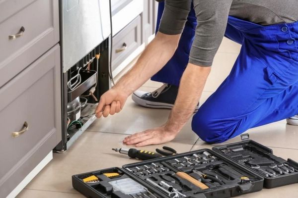 A repairman in blue overalls crouches beside a kitchen appliance, using tools from an open toolbox