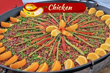Large pan of mild chicken paella garnished with asparagus, red peppers, peas, and orange slices.