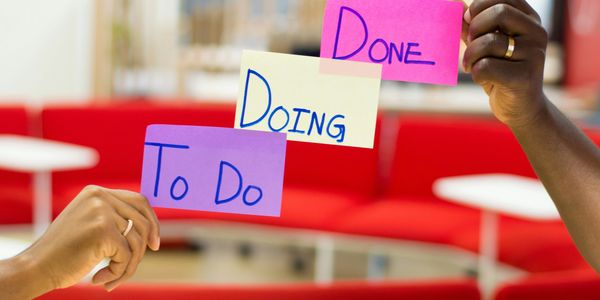 Hands holding sticky notes labeled To Do, Doing, and Done against a red couch background.