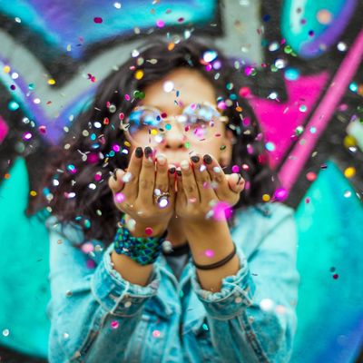 Person blowing colorful confetti in front of graffiti wall.