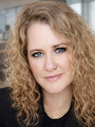 Portrait of a woman with curly blonde hair and blue eyes, wearing a nose ring and necklace.