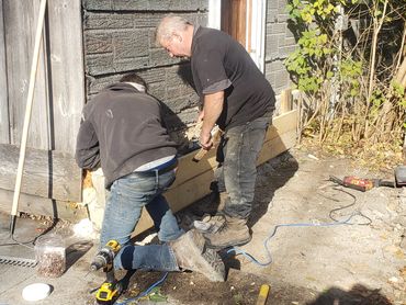 Two men working on a wooden foundation outside a building.