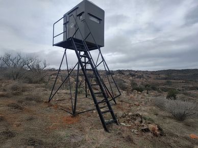 A black hunting blind elevated on metal legs in a dry, rocky landscape under a cloudy sky.