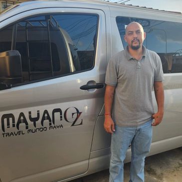 Man standing beside a silver van with Mayan travel logos.