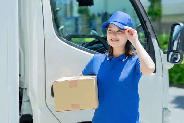 A smiling delivery woman in a blue uniform and cap holds a package next to a white van