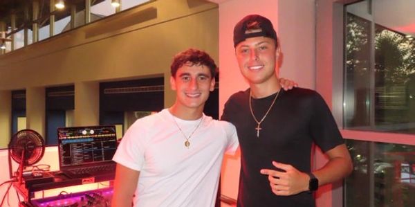 Two young men smiling, posing indoors near DJ equipment with warm lighting.