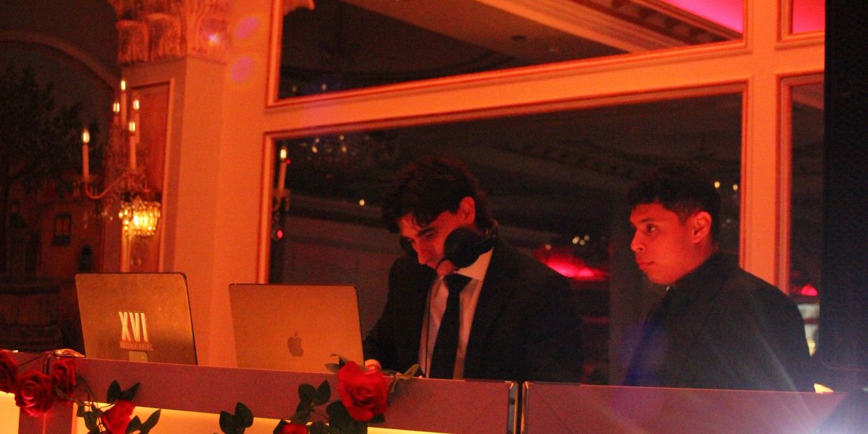 Two men DJing with laptops in dim, warm lighting.