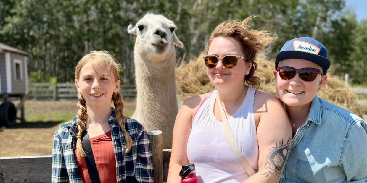 Three people posing with a llama outdoors on a sunny day.