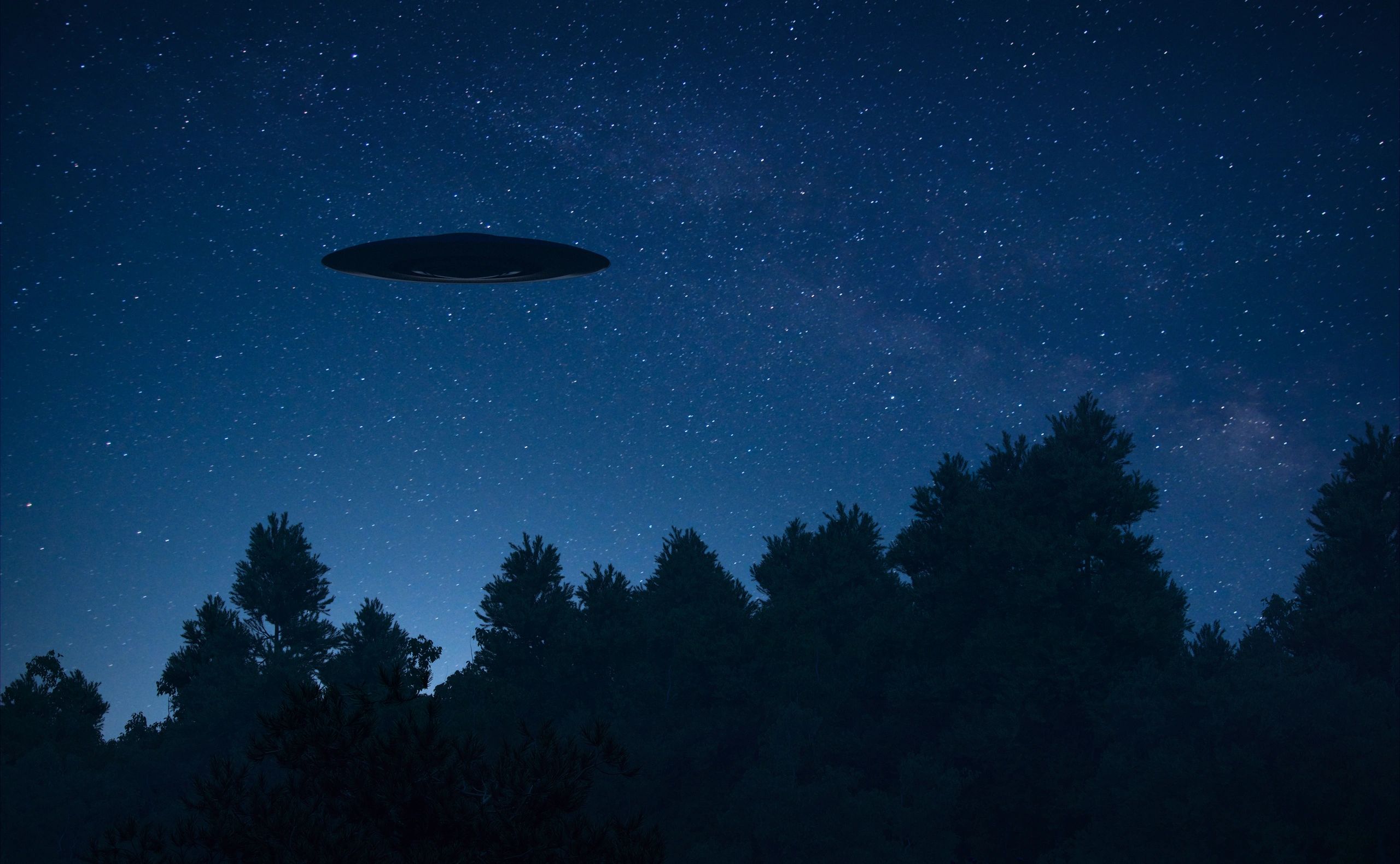 Saucer silouette hovers over trees against a backdrop of stars in the night sky.
