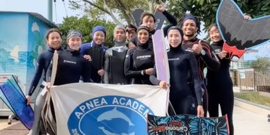 A group of divers in wetsuits holding an Apnea Academy banner and monofins outdoors.