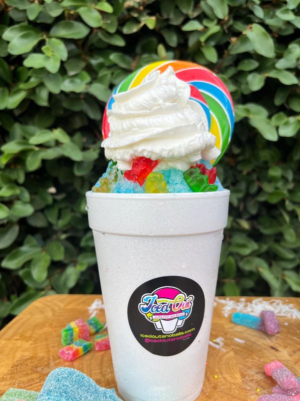 Colorful shaved ice dessert topped with whipped cream, gummy bears, and a rainbow lollipop.