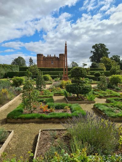 A historic castle behind a manicured garden with a tall sundial under a partly cloudy sky.