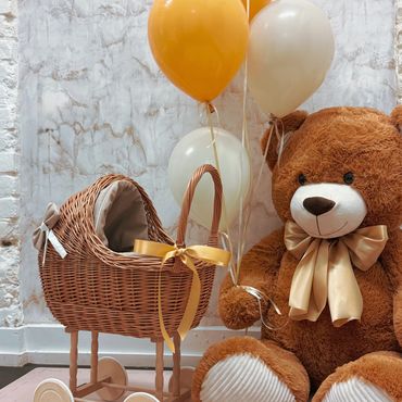 A teddy bear holding balloons next to a wicker baby carriage with a yellow bow.