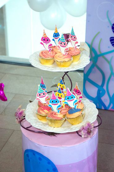 Cupcakes with colorful Baby Shark toppers on a two-tier stand at a party.