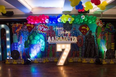 Colorful superhero-themed birthday backdrop with balloons and illuminated number 7.
