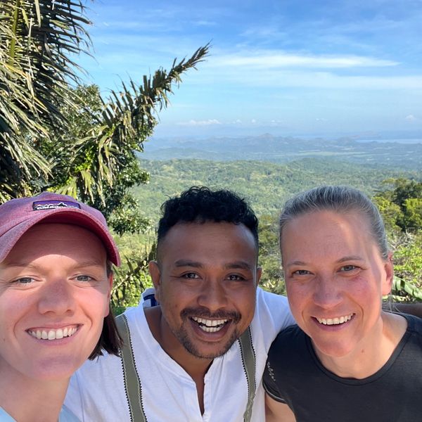 Three friends smiling for a selfie with a scenic mountain and sky background.