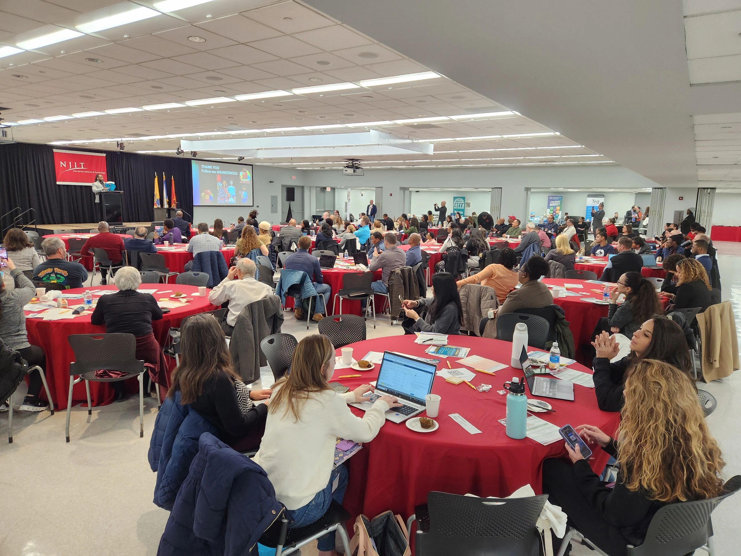 A large audience attentively listens to a speaker at a conference with red-covered tables.