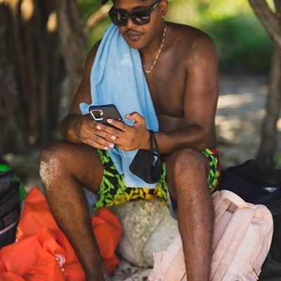 Man in sunglasses using phone on beach with towel and bags.