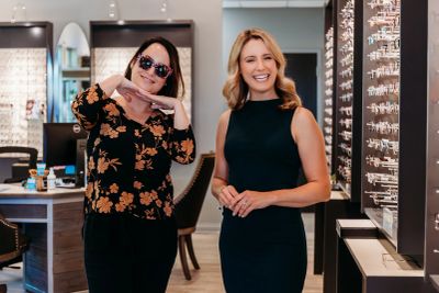 Two women smile in an optical store, one wearing sunglasses.
