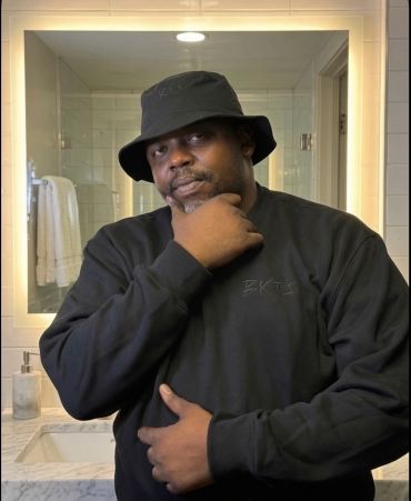 Man in black hoodie and bucket hat posing in front of a bathroom mirror.