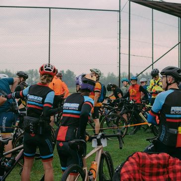 Cyclists gather before a race at a fenced sports field.