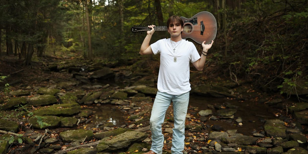 Man standing in forest with guitar over shoulder.
