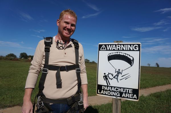 Smiling man in parachute gear near a warning sign at parachute landing area.