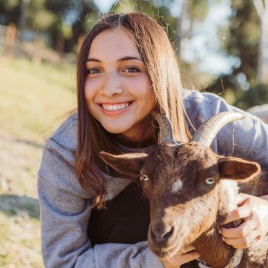 A young woman smiles while hugging a brown goat outdoors.