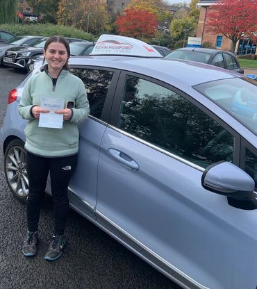 A smiling woman proudly holding her driving test pass certificate next to a driving school car.