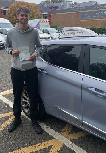 Young man holding a driving test pass certificate next to a driving school car.