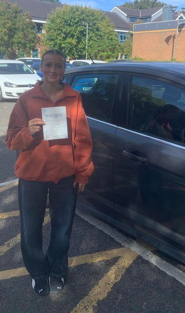 Young woman proudly holding her practical driving test pass certificate beside a car.
