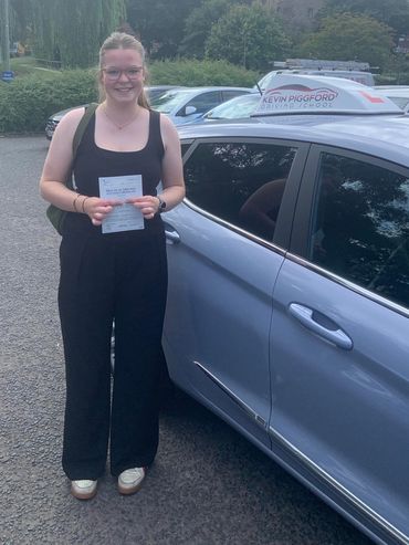A smiling young woman holds her practical driving test pass certificate next to a driving school car.