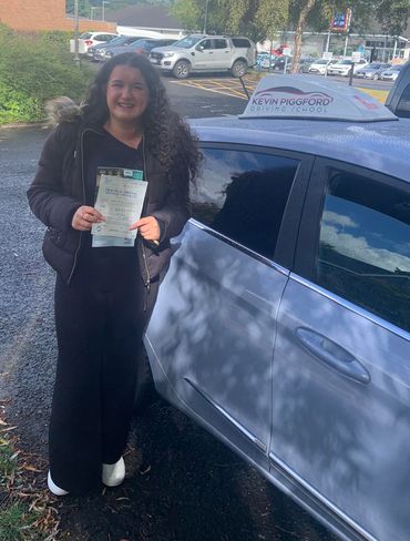 Young woman proudly holding her driving test pass certificate beside a driving school car.