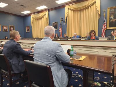 Officials in a formal hearing or meeting room with American flags.