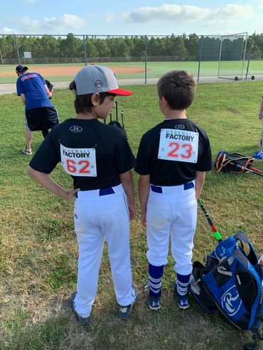 Two young baseball players in uniforms with numbers 62 and 23 stand on grass near a baseball field.