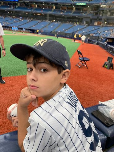 Young boy in a Yankees cap and jersey holding a baseball at a stadium.