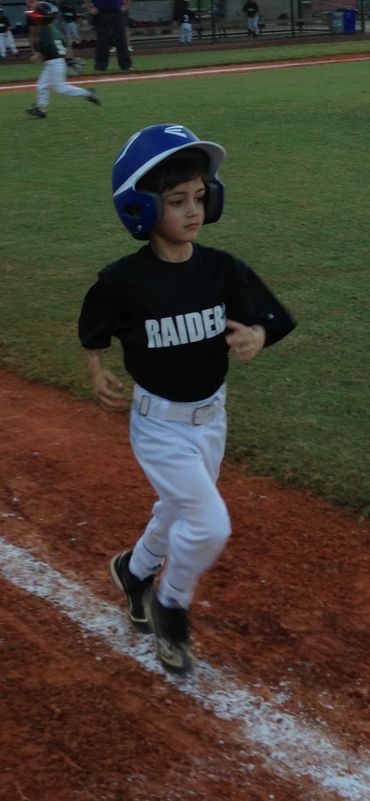 Young baseball player running on the field wearing a blue helmet and Raiders jersey.