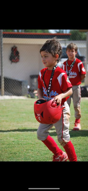 Young boy in baseball uniform holding a red helmet with a second boy in the background.