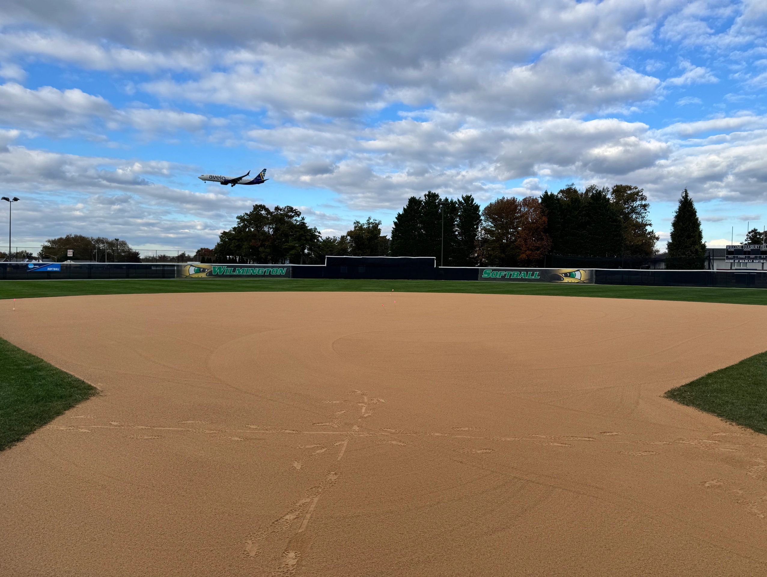 A softball field with an airplane flying in the sky above.