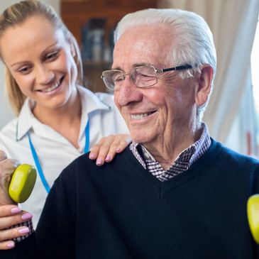 Elderly man exercising with dumbbells assisted by a smiling caregiver.