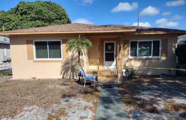 Small beige house with two windows, a front door, and a palm tree casting shadow.