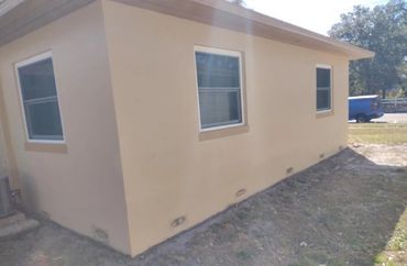 Side view of a beige house with three windows and dry grass around it.