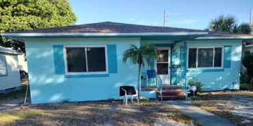 A small turquoise house with a palm tree and two chairs in the front yard.