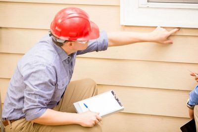 Home inspector examining house exterior with clipboard and red helmet.