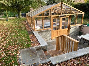 Wood and glass greenhouse with cozy seating inside in a leafy garden.