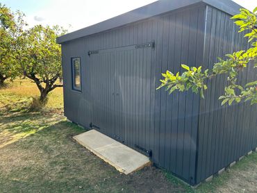 A modern grey garden shed with double doors and a small window surrounded by trees.