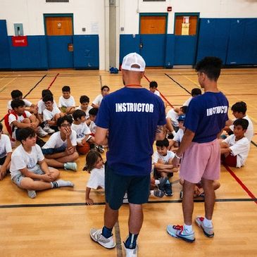 Three instructors talk to children sitting on a gym floor in a basketball setting.