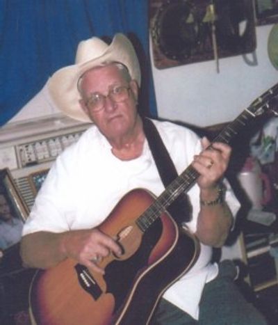 Elderly man in a cowboy hat playing an acoustic guitar indoors.