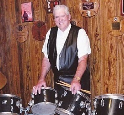Elderly man in a leather vest smiling behind a drum set.