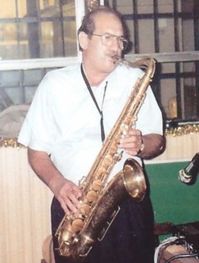 Man playing a saxophone indoors, wearing glasses and a white shirt.